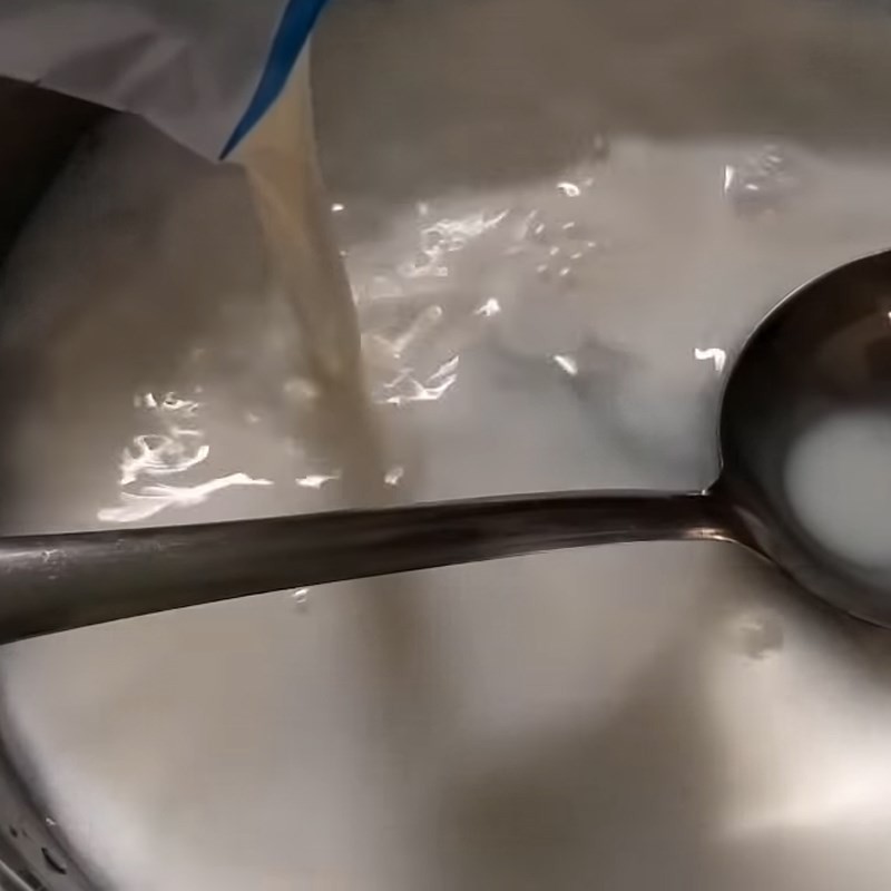 Một người đang dùng muỗng khuấy đều hỗn hợp bột rau câu, đường, sữa tươi và nước lọc trong nồi trên bếp, hỗn hợp có màu trắng đục