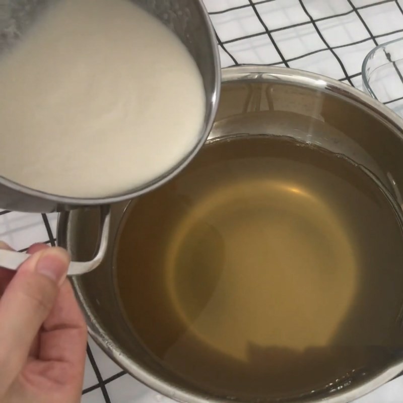 Một người đang dùng muỗng khuấy đều hỗn hợp nước cốt dừa và rau câu trong một chiếc nồi lớn, tạo thành chất lỏng màu trắng ngà